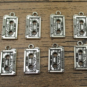 Puede incluir: Ocho abalorios de metal plateados en forma de casetes. Cada abalorio tiene un diseño detallado de un casete con un patrón en blanco y negro.