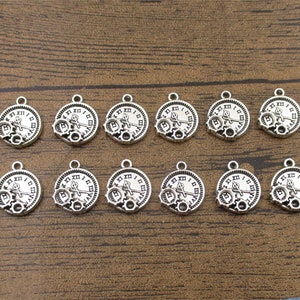 Könnte beinhalten: Eine Sammlung silberfarbener Uhr-Anhänger mit römischen Ziffern und Zahnrad-Details. Jeder Anhänger hat eine kleine Schlaufe zum Befestigen an Schmuck oder anderen Bastelarbeiten. Die Anhänger liegen auf einer braunen Holzoberfläche.