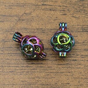 以下が含まれることがあります： 丸い形をした、虹色の金属製のチャームケージが2つ。複雑なデザインが施されています。ケージは片側が開いており、上部に吊り下げ用のループが付いています。