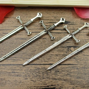 May include: Four silver-toned sword-shaped keychains with intricate details. The keychains are arranged in a row on a wooden surface.