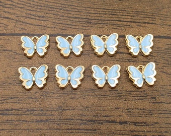 May include: Eight butterfly charms with a light blue enamel finish and gold-colored metal edges. The charms are arranged on a brown wooden surface, ideal for jewelry making or crafting.