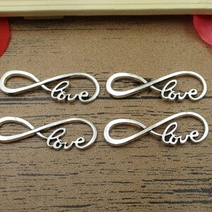 May include: Four silver infinity symbol charms with the word "love" inscribed on each charm. The charms are on a wooden surface.