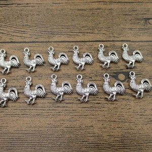 May include: A collection of 14 silver-toned rooster charms, each with a detailed design. The charms are arranged in two rows on a wooden surface.