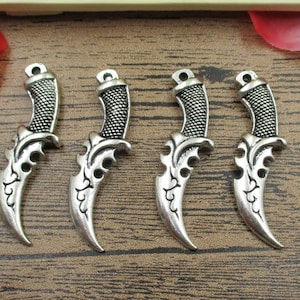Peut inclure: Quatre breloques en forme de poignard en métal argenté avec des détails complexes. Les breloques sont en forme de poignard avec une lame incurvée et une poignée décorative.