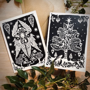 K&ouml;nnte beinhalten: Zwei Schwarz-Wei&szlig;-Kunstdrucke mit detaillierten Linolschnitt-Designs. Einer zeigt einen Engel mit Fl&uuml;geln, der andere einen Baum mit dem Wort "Yule". Beide sind auf cremefarbenem Papier, mit einem Kraftpapierumschlag im Hintergrund.