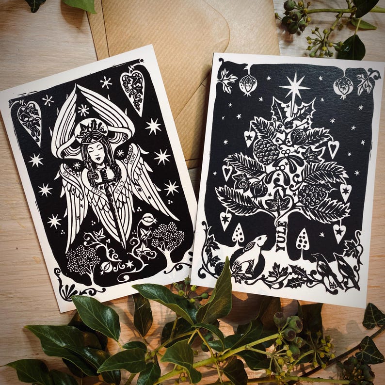 K&ouml;nnte beinhalten: Zwei Schwarz-Wei&szlig;-Kunstdrucke mit detaillierten Linolschnitt-Designs. Einer zeigt einen Engel mit Fl&uuml;geln, der andere einen Baum mit dem Wort "Yule". Beide sind auf cremefarbenem Papier, mit einem Kraftpapierumschlag im Hintergrund.