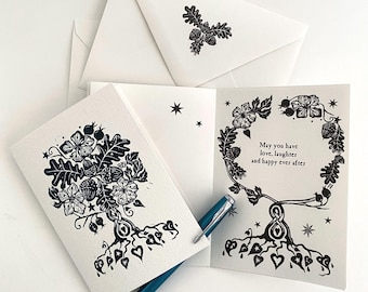 Könnte beinhalten: Eine weiße Grußkarte mit einer schwarzen Tuschezeichnung eines Baumes mit Eichenblättern, Eicheln und herzförmigen Wurzeln. Der Innentext lautet: "May you have love, laughter and happy ever after". Ein blauer Stift liegt auf der Karte, daneben ein Umschlag.