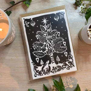 K&ouml;nnte beinhalten: Schwarz-wei&szlig;e Weihnachtskarte mit einem Baumdesign, Sternen, Beeren und dem Wort "Yule". Die Karte befindet sich in einer transparenten Plastikh&uuml;lle mit einem braunen Papierr&uuml;cken. Gr&uuml;ne Bl&auml;tter und eine brennende Kerze umgeben die Karte.