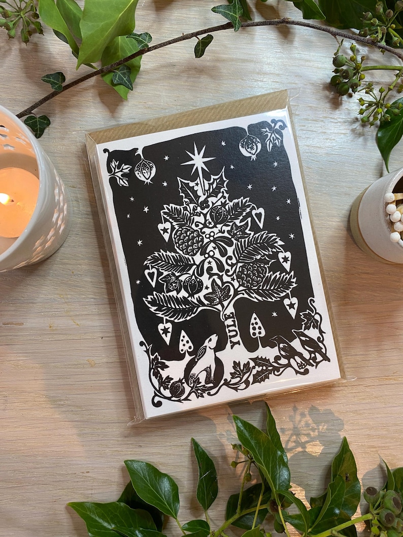 K&ouml;nnte beinhalten: Schwarz-wei&szlig;e Weihnachtskarte mit einem Baumdesign, Sternen, Beeren und dem Wort "Yule". Die Karte befindet sich in einer transparenten Plastikh&uuml;lle mit einem braunen Papierr&uuml;cken. Gr&uuml;ne Bl&auml;tter und eine brennende Kerze umgeben die Karte.