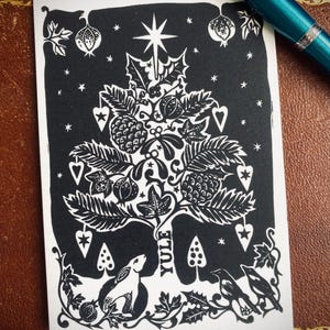 K&ouml;nnte beinhalten: Schwarz-wei&szlig;e Yule-Karte mit einem stilisierten Weihnachtsbaum-Design. Die Karte zeigt einen Stern, Bl&auml;tter, Beeren und das Wort "Yule". Ein Hase und V&ouml;gel befinden sich am Fu&szlig;e des Baumes. Ein t&uuml;rkisfarbener Stift liegt auf der braunen Oberfl&auml;che.