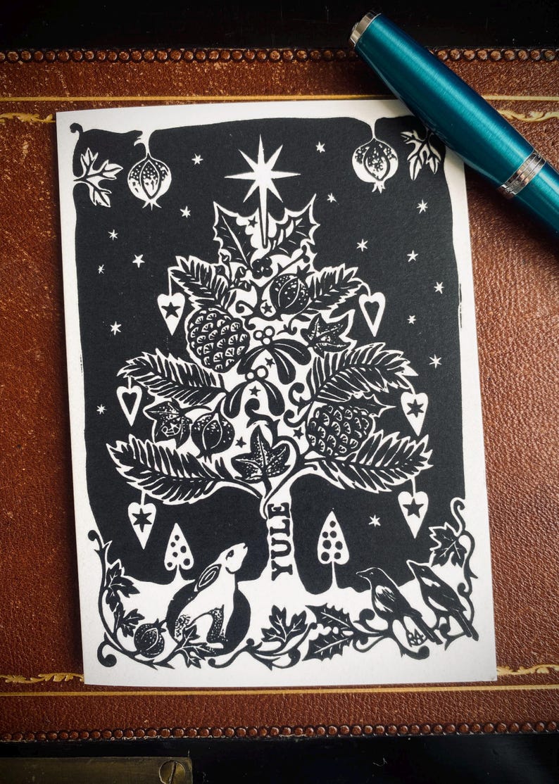 K&ouml;nnte beinhalten: Schwarz-wei&szlig;e Yule-Karte mit einem stilisierten Weihnachtsbaum-Design. Die Karte zeigt einen Stern, Bl&auml;tter, Beeren und das Wort "Yule". Ein Hase und V&ouml;gel befinden sich am Fu&szlig;e des Baumes. Ein t&uuml;rkisfarbener Stift liegt auf der braunen Oberfl&auml;che.