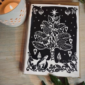 K&ouml;nnte beinhalten: Ein Stapel schwarz-wei&szlig;er Yule-Karten mit einem detaillierten Baumdesign, einem Stern, Beeren und dem Wort "Yule". Die Karten befinden sich in einer transparenten Plastikh&uuml;lle, mit einer brennenden Kerze und Gr&uuml;nzeug im Hintergrund.