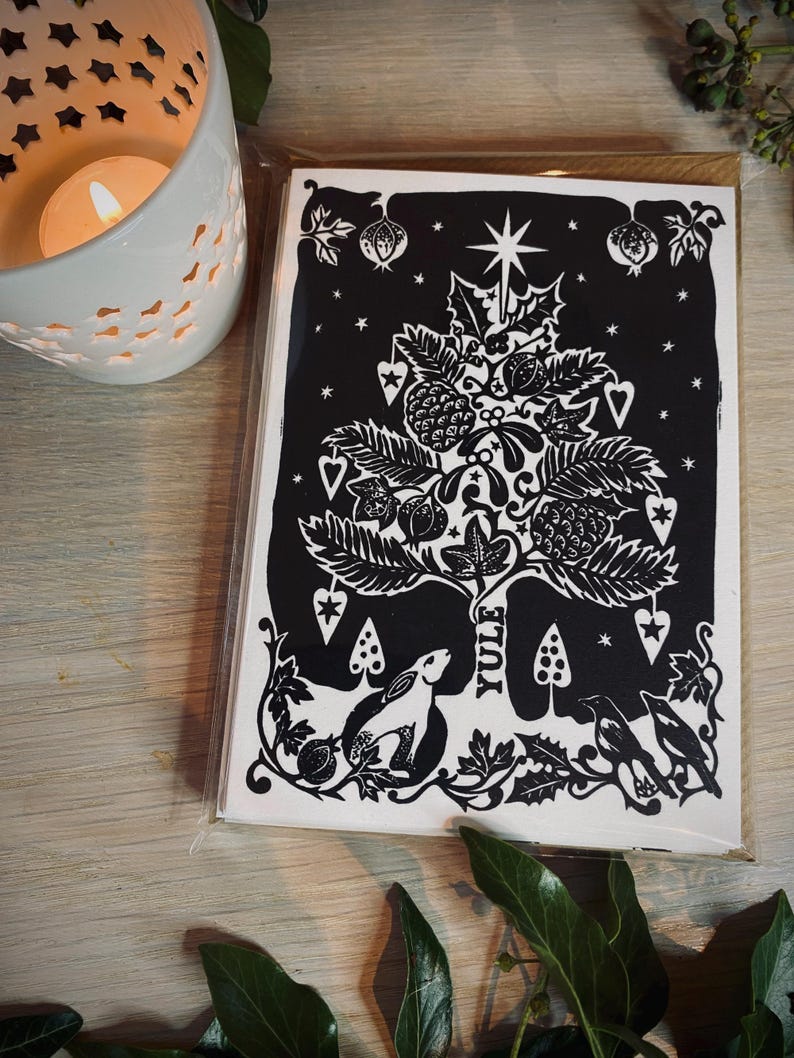 K&ouml;nnte beinhalten: Ein Stapel schwarz-wei&szlig;er Yule-Karten mit einem detaillierten Baumdesign, einem Stern, Beeren und dem Wort "Yule". Die Karten befinden sich in einer transparenten Plastikh&uuml;lle, mit einer brennenden Kerze und Gr&uuml;nzeug im Hintergrund.