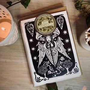 K&ouml;nnte beinhalten: Ein Stapel schwarz-wei&szlig;er Kunstkarten mit einem Engel-Design und den Worten "Joy, Love, Peace, Hope". Die Karten sind in klarem Kunststoff mit einem Raw Alchemy-Aufkleber versiegelt. Eine brennende Kerze und Streichh&ouml;lzer sind im Hintergrund.