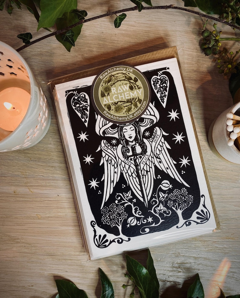 K&ouml;nnte beinhalten: Ein Stapel schwarz-wei&szlig;er Kunstkarten mit einem Engel-Design und den Worten "Joy, Love, Peace, Hope". Die Karten sind in klarem Kunststoff mit einem Raw Alchemy-Aufkleber versiegelt. Eine brennende Kerze und Streichh&ouml;lzer sind im Hintergrund.