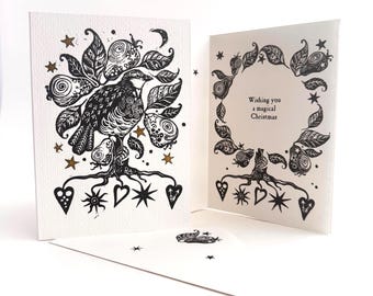Könnte beinhalten: Eine weiße Grußkarte mit einer schwarzen Tuschezeichnung eines Vogels auf einem Baum mit Birnen, Sternen und Herzen. Der Innentext lautet, Wishing you a magical Christmas. Ein passender Umschlag mit einer kleinen Vogelillustration liegt bei.