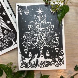 K&ouml;nnte beinhalten: Schwarz-wei&szlig;e Yule-Gru&szlig;karte mit einem stilisierten Weihnachtsbaum-Design. Der Baum ist mit Sternen, Herzen und anderen festlichen Elementen geschm&uuml;ckt. Ein Hase und V&ouml;gel befinden sich am Fu&szlig;e des Baumes. Das Wort "YULE" ist vertikal gedruckt.