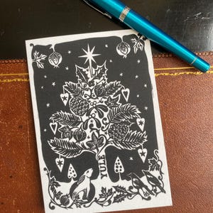 K&ouml;nnte beinhalten: Schwarz-wei&szlig;e Weihnachtskarte mit einem Yule-Baum-Design, mit einem Stern, Beeren und einem Hasen. Ein t&uuml;rkisfarbener Stift liegt auf der Karte. Die Karte liegt auf einer braunen Oberfl&auml;che.