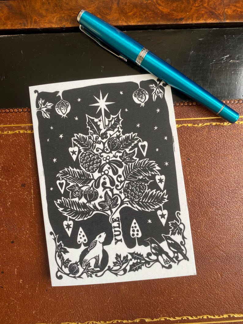 K&ouml;nnte beinhalten: Schwarz-wei&szlig;e Weihnachtskarte mit einem Yule-Baum-Design, mit einem Stern, Beeren und einem Hasen. Ein t&uuml;rkisfarbener Stift liegt auf der Karte. Die Karte liegt auf einer braunen Oberfl&auml;che.