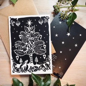 K&ouml;nnte beinhalten: Eine schwarz-wei&szlig;e Weihnachtskarte mit einem Baumdesign, das einen Stern, Herzen und andere festliche Elemente zeigt. Das Wort "Yule" ist auf die Karte gedruckt. Ein brauner Umschlag und eine schwarze Karte mit wei&szlig;en Sternen sind ebenfalls zu sehen.