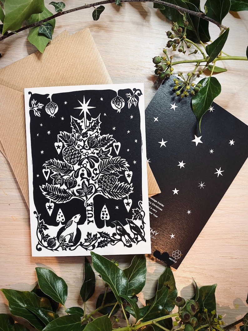 K&ouml;nnte beinhalten: Eine schwarz-wei&szlig;e Weihnachtskarte mit einem Baumdesign, das einen Stern, Herzen und andere festliche Elemente zeigt. Das Wort "Yule" ist auf die Karte gedruckt. Ein brauner Umschlag und eine schwarze Karte mit wei&szlig;en Sternen sind ebenfalls zu sehen.