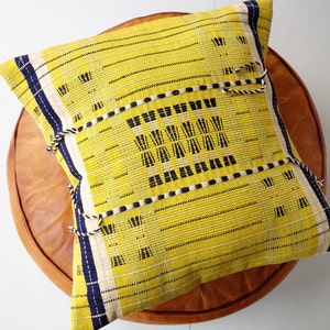 May include: A square yellow decorative pillow with a woven design, featuring navy blue and white stripes and black geometric patterns. The pillow is adorned with decorative rope tassels and rests on a brown leather ottoman.