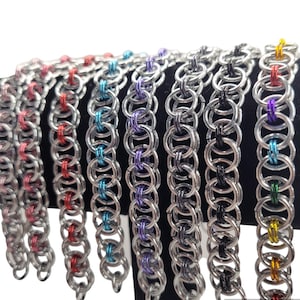May include: A collection of chainmail bracelets in various colors. The bracelets are made of silver-colored metal rings, with some rings in red, blue, purple, black, and yellow. The bracelets are displayed against a black background.