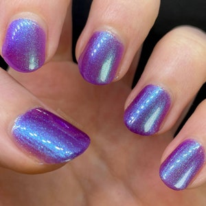 May include: Close-up of five fingernails painted with a holographic purple nail polish. The polish has a blue and green iridescent shimmer.