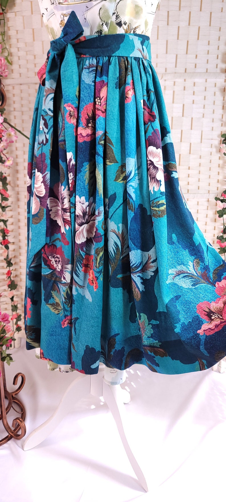 May include: A teal skirt with a floral pattern featuring large pink, white, and purple flowers. The skirt has a wide waistband with a matching tie and is displayed on a white dress form. The fabric has a corduroy texture.