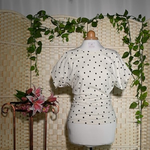 Pode incluir: Uma blusa branca com bolinhas pretas e mangas curtas. A blusa é ajustada e tem um decote redondo.