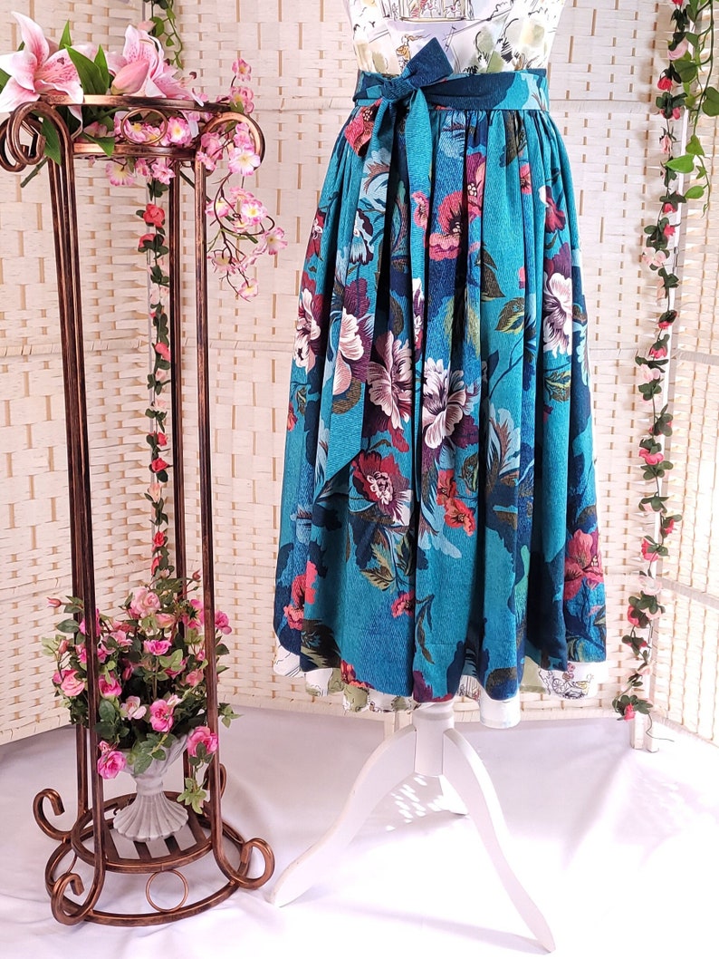 May include: A teal floral skirt with a wide waistband and a matching tie. The skirt features a vibrant pattern of large flowers in shades of pink, red, and purple. The skirt is displayed on a white dress form.