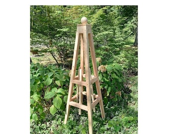 May include: A tall, wooden garden obelisk with a square base and a pointed top. The obelisk is made of unfinished wood and has a decorative ball finial. It stands in a garden setting with green foliage in the background.