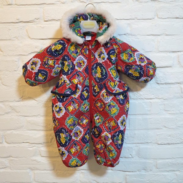 May include: A red, long-sleeved, one-piece snowsuit featuring a repeating pattern of yellow cartoon bears in blue and green frames. The garment includes a front zip closure, two front pockets with flaps, and a hood trimmed with white faux fur.