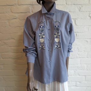 Pode incluir: Uma camisa de manga comprida com botões num padrão de xadrez vichy azul e branco. A parte frontal apresenta dois painéis verticais de desenhos bordados, incluindo pequenos ursos de peluche, corações axadrezados e motivos florais.