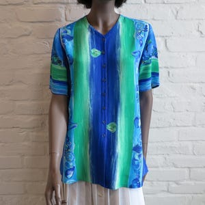 Op de afbeelding: Een overhemd met knopen en een V-hals, met verticale blauwe en groene strepen. Het shirt heeft korte mouwen en een blauw bloemmotief. Het kledingstuk wordt over een witte rok gedragen.