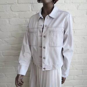 May include: A pale purple denim jacket with a button-up front, two chest pockets, and a collared neckline. The jacket has visible stitching and is paired with a white pleated skirt. The jacket appears to be a size medium.