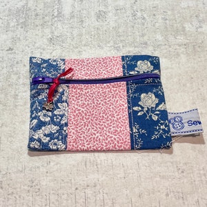 May include: A small blue and pink zippered pouch with a floral pattern. The pouch has a purple zipper and a red ribbon with a silver charm. The pouch has a white label with the word "Sew" on it.