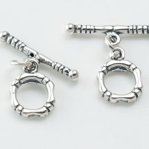 Op de afbeelding: Twee zilveren toggle-sluitingen met een ronde, getextureerde ring die aan elk is bevestigd. De ringen hebben een licht verhoogd, cirkelvormig ontwerp.