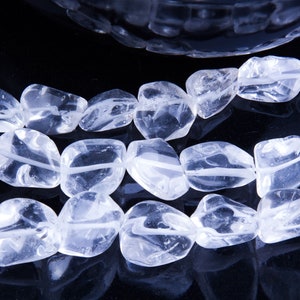 Pode incluir: Um fio de contas de cristal de quartzo transparente, cada conta tem uma forma irregular e lisa. As contas est&atilde;o dispostas em uma fila sobre uma superf&iacute;cie preta.