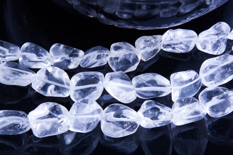 Pode incluir: Um fio de contas de cristal de quartzo transparente, cada conta tem uma forma irregular e lisa. As contas est&atilde;o dispostas em uma fila sobre uma superf&iacute;cie preta.
