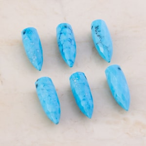 May include: Six turquoise blue gemstone bullet-shaped beads with a pointed tip. The beads are smooth and have a translucent appearance.