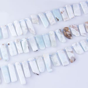 May include: A strand of light blue and white gemstone beads, each bead is a long, irregular rectangle shape. The beads are strung on a thin, white thread.