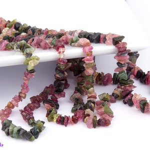 May include: A strand of multi-colored, chip-shaped, natural gemstone beads. The beads are pink, green, and black. The beads are strung on a clear thread.