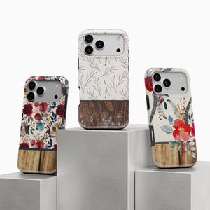 May include: Three phone cases with unique designs. One case features a floral pattern with red and teal flowers, another has a white background with brown branches, and the third showcases a floral and antler design. Each case has a wooden base.