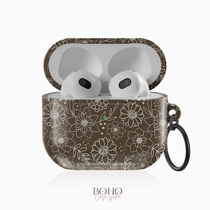 Op de afbeelding: Open bruine AirPods-hoes met bloemmotieven en witte oortelefoons erin. De hoes heeft een wit bloemmotief en een zwarte ring voor bevestiging. De tekst "BOHO Case studio" staat onderaan.