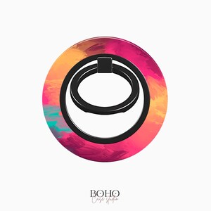 May include: A round phone ring holder with a black ring and a colorful abstract design. The design features shades of pink, orange, and blue.