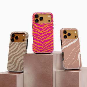 May include: Three phone cases with unique designs. One case features a vibrant pink and orange zebra stripe pattern. The other two cases have a neutral color palette with abstract wave and zebra stripe designs. All cases have a gold-colored camera housing.