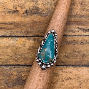 May include: A teardrop-shaped turquoise stone ring set in a silver bezel. The setting features a twisted rope border and small silver bead accents along the edges. The stone displays a vibrant teal color with natural brown matrix inclusions.