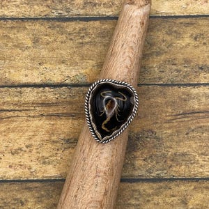 Op de afbeelding: Een hartvormige ring met een zwart midden met een schorpioenontwerp. De ring heeft een zilverkleurige, getextureerde rand. De ring wordt getoond op een houten deuvel tegen een houten achtergrond.