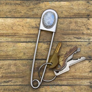 Pode incluir: Um porta-chaves grande em forma de alfinete de ama prateado com uma pedra da lua oval engastada numa borda de metal torcido no topo. Um anel de metal circular está preso à base do alfinete, segurando um conjunto de três chaves de metal.